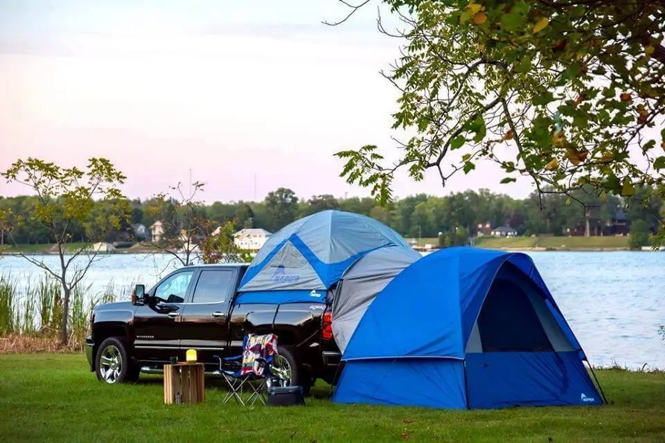 Truck bed camping with a truck on a green grass near river with blue tent on the back and tent on the ground