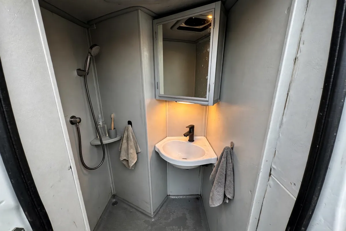 Truck camper wet bath with a small corner sink, mirror, and medicine cabinet under warm LED light