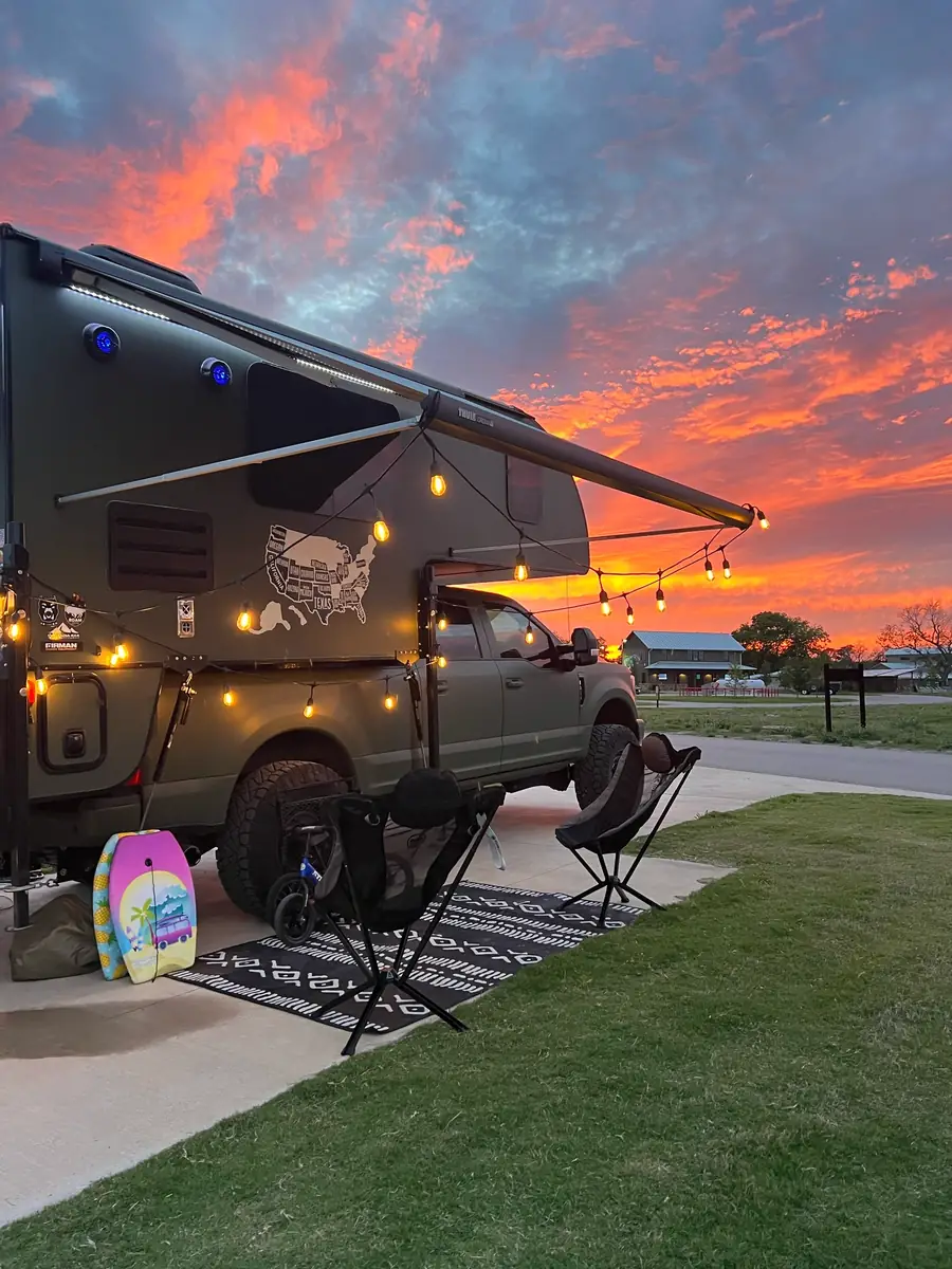 Truck camper set up as a home base with awning extended, chairs and outdoor rug at sunset