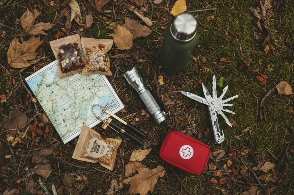 Flat lay of camping safety essentials including a first-aid kit, flashlight, multi-tool, and map on the forest floor.